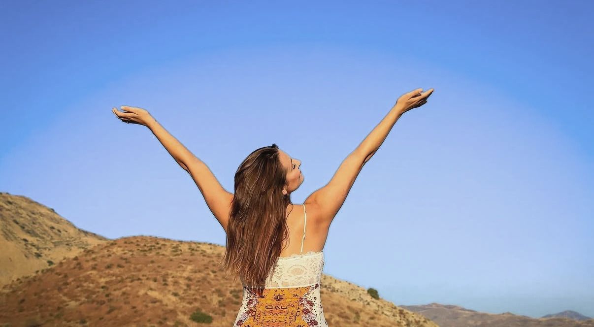 Cara on a sunlit hillside with her back to the camera and her arms raised to the sky.