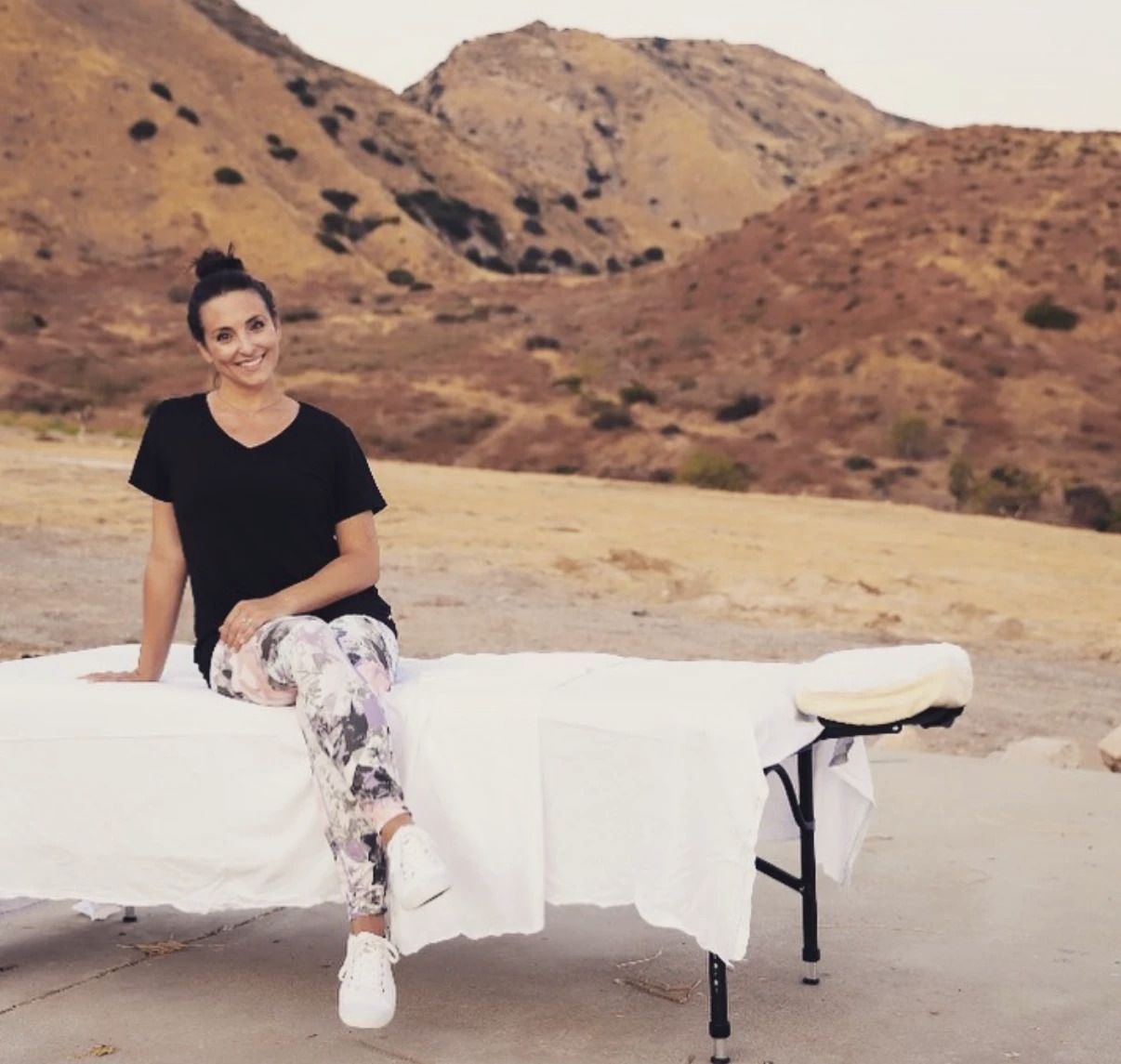 Cara smiling, sitting on her massage table outdoors in the golden hills outside Los Angeles.